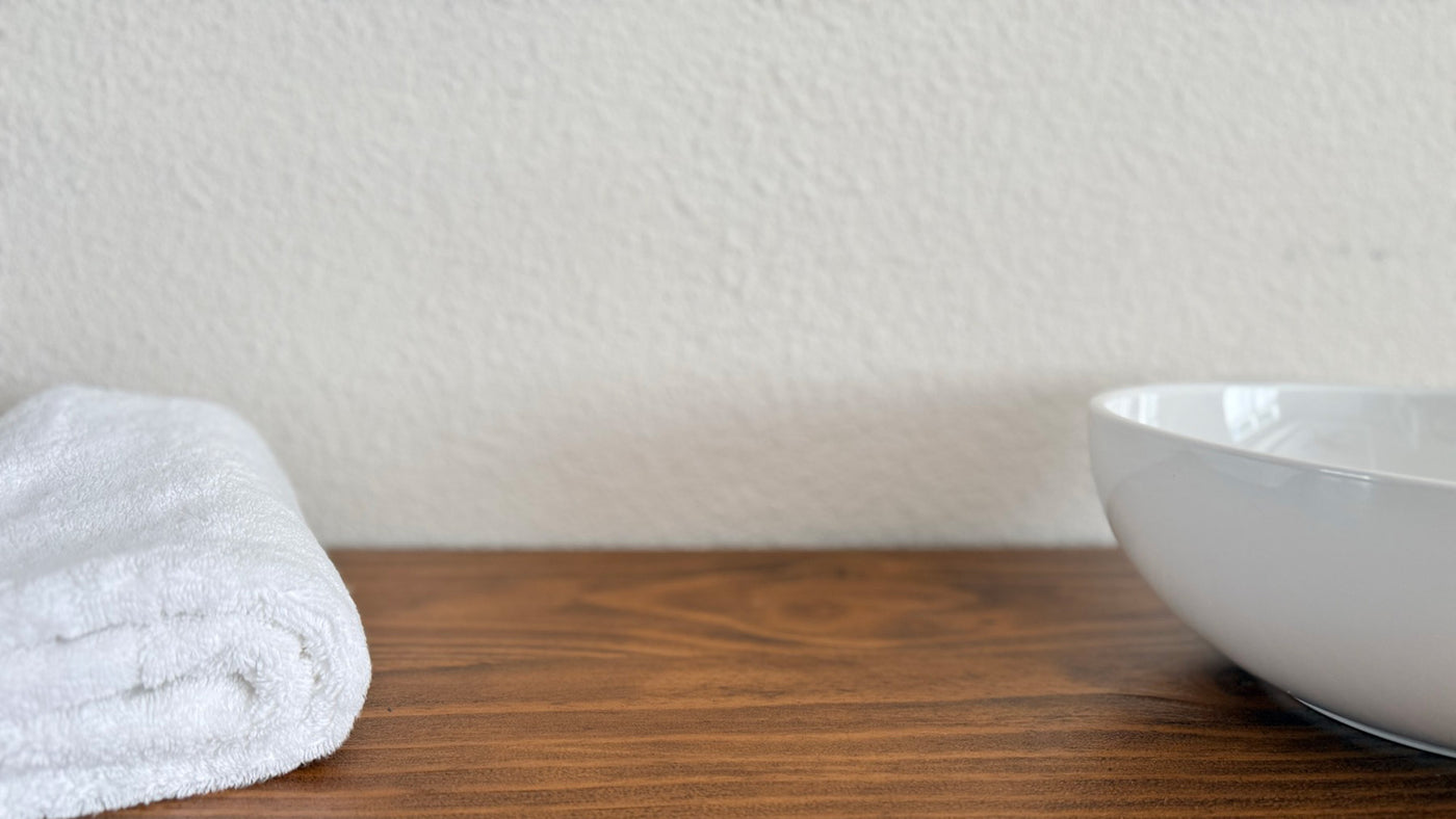Wellua hero, desktop. A small ceramic mixing bowl and folded warm-cream towel on a stucco shelf in honest light, leaving the upper two-thirds open for the headline.